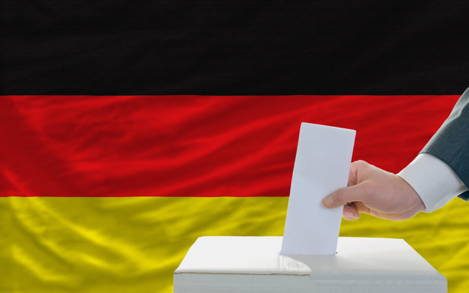 Eine Hand wirft einen Zettel in eine Wahlurne. Im Hintergrund die Fahne der Bundesrepublik Deutschland.